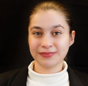 Portrait of Margi who is a young woman with a medium light skin tone and medium brown hair pulled back behind her head. She has a close-mouthed smile and wears a white turtleneck sweater under a black suit jacket.