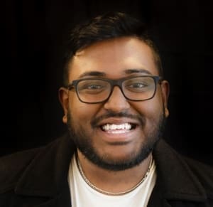 A portrait of Naflan, who is a young man with a medium-dark skin tone, short dark hair, and a dark beard and moustache. He smiles toothily and wears black-rimmed glasses with a white t-shirt and lack cardigan sweater.